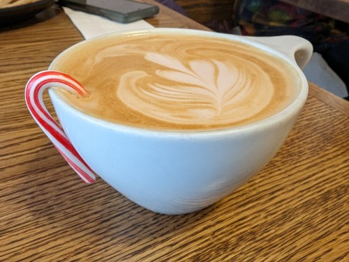 A white cup of latte art topped with a heart design, featuring a small candy cane on the rim, placed on a wooden table.