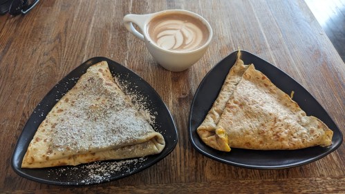 Two triangular crepes on black plates, one dusted with powdered sugar, alongside a cup of latte with a swirl design.