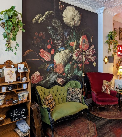 A cozy interior featuring a floral mural, a green tufted sofa, a red armchair, and a wooden shelf with decor items.