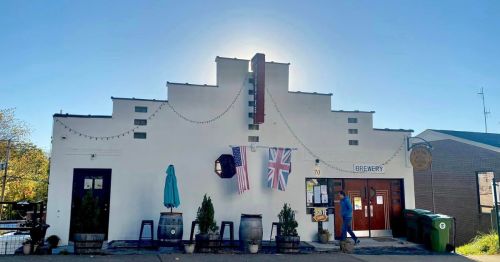 A brewery building with flags, outdoor seating, and a clear blue sky in the background.