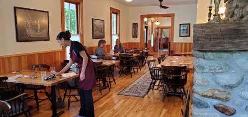 A cozy restaurant interior with wooden tables, a server setting a table, and diners in a warm, inviting atmosphere.