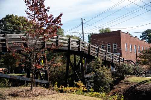 A wooden bridge spans a small creek, with a brick building and trees in the background under a clear blue sky.