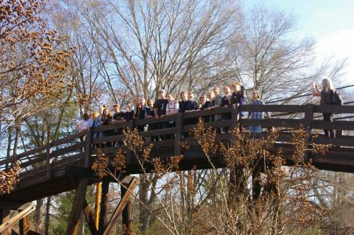 A group of people stands on a wooden bridge surrounded by trees, smiling and posing for a photo.