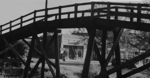 A wooden bridge with a support structure, leading to a building in the background, captured in black and white.