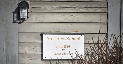 Sign on a weathered wooden wall reads "North St. School, built 1866 by Jacob Stewart," with a lantern above.