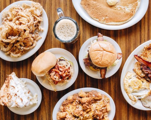 A spread of various comfort foods including burgers, fried chicken, pancakes, onion rings, and a mug of beer.