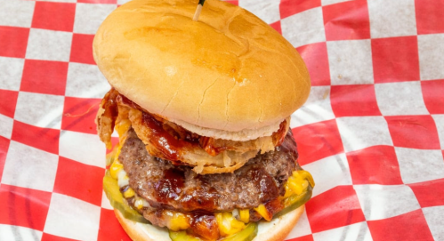 A stacked burger with two patties, cheese, pickles, crispy onions, and barbecue sauce on a red and white checkered plate.