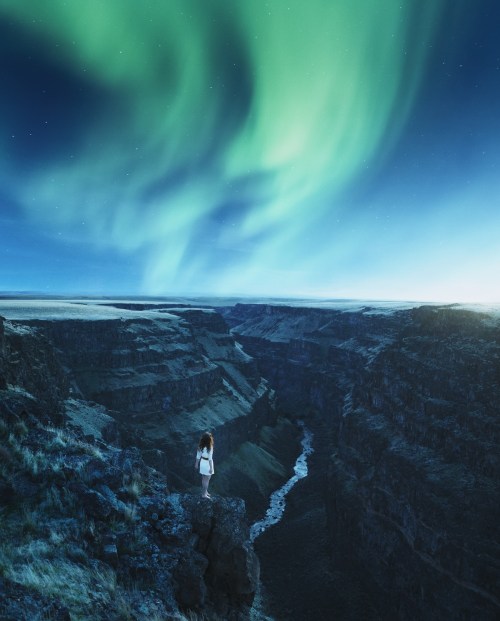 A person stands on a cliff overlooking a canyon, illuminated by vibrant northern lights in the night sky.