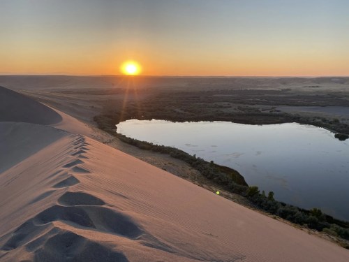 Sunset over a serene lake, with sand dunes in the foreground and a vast landscape stretching into the distance.