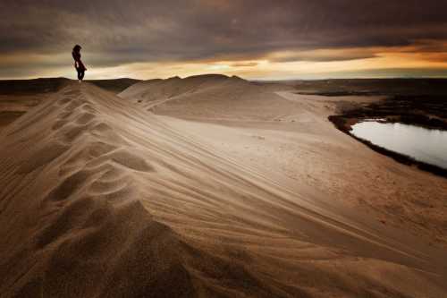 A person stands on a sandy dune at sunset, overlooking a river and a vast landscape under a dramatic sky.