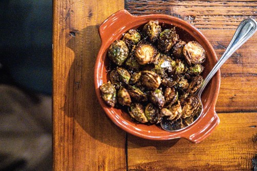 A bowl of roasted Brussels sprouts on a wooden table, with a spoon resting inside the dish.