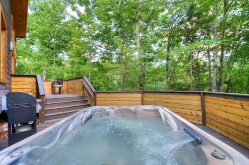 A hot tub on a wooden deck surrounded by lush green trees, with a grill and seating area nearby.