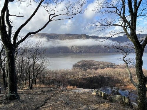 A serene landscape featuring bare trees, a calm lake, and misty mountains in the background under a cloudy sky.