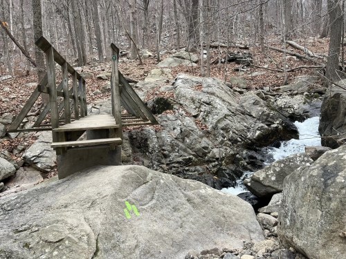 A wooden bridge crosses a rocky stream in a forested area with bare trees and scattered leaves.