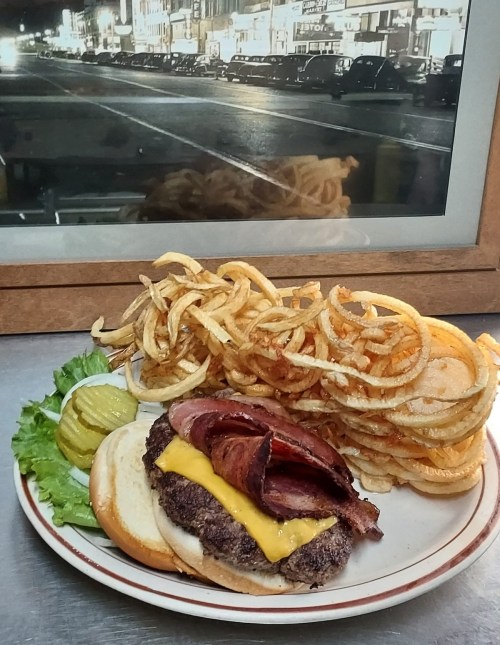 A cheeseburger topped with bacon, served with curly fries and pickles, with a vintage city scene in the background.