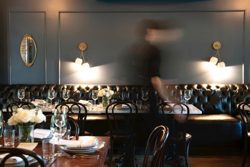 A blurred figure of a server moves through a stylish restaurant with dark blue walls and elegant table settings.