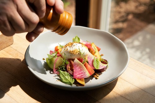 A hand sprinkles pepper over a colorful salad with burrata, tomatoes, and fresh herbs on a light plate.
