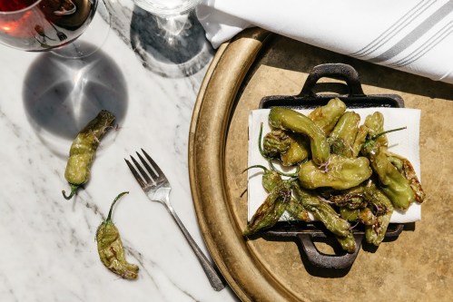 A plate of grilled shishito peppers on a marble table, accompanied by a fork and a glass of wine.