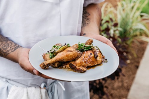 A person holds a plate with two roasted chicken legs topped with greens and garnishes.