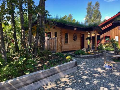 A cozy wooden building with a green roof, surrounded by trees and colorful flowers in a garden setting.