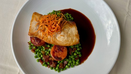 A plated dish featuring a crispy fish fillet on a bed of green peas, with sliced sausage and a dark sauce.