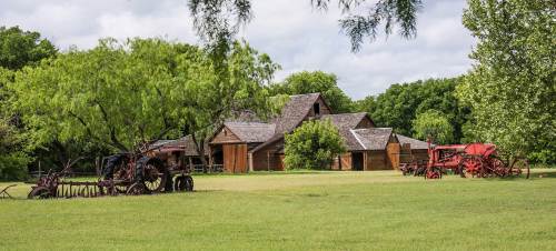 Rustic farm scene featuring vintage tractors and wooden buildings surrounded by lush greenery under a cloudy sky.