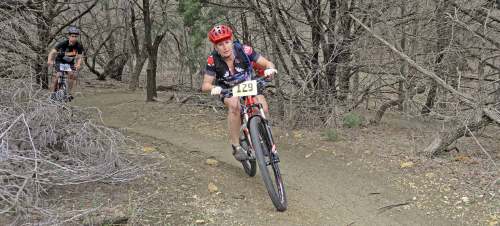 A mountain biker navigates a dirt trail surrounded by trees, with another cyclist in the background.