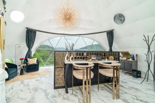 A stylish interior of a geodesic dome featuring a bar area, elegant chairs, and scenic views through large windows.