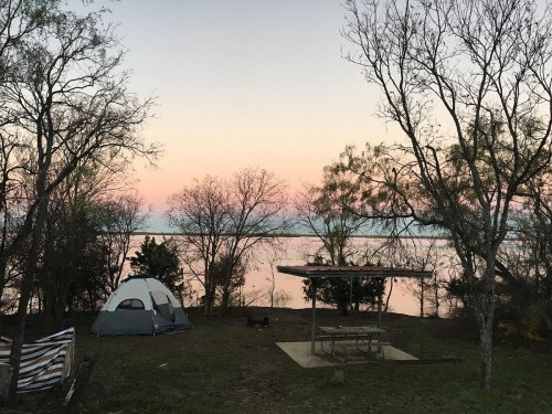 A serene campsite by a lake at sunset, featuring a tent and picnic area surrounded by trees.