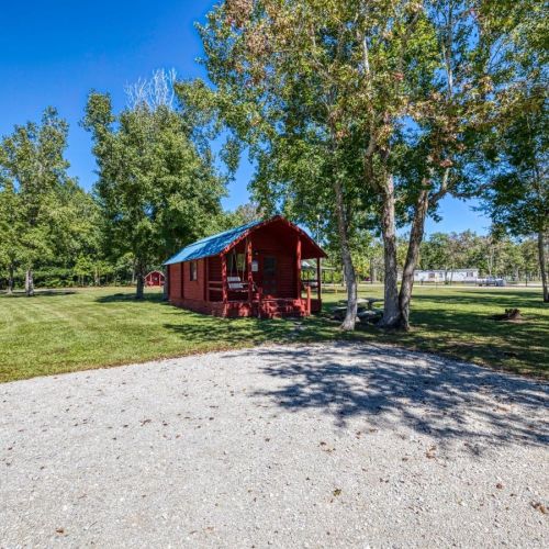 A red cabin with a blue roof surrounded by trees and a grassy area on a sunny day.
