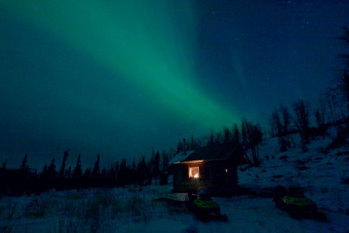 A cozy cabin illuminated at night under a vibrant green aurora borealis in a snowy landscape.