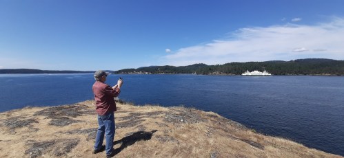 A person stands on a rocky shore, taking a photo of a ferry on the water with a forested landscape in the background.
