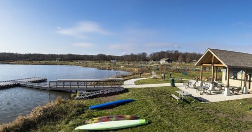 A serene lakeside view with kayaks, a dock, and a picnic area under a clear blue sky.