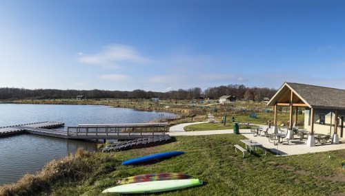 A serene lakeside view with a dock, colorful kayaks, and a picnic area under a clear blue sky.