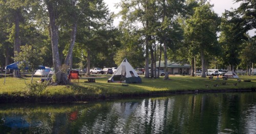 A serene campsite by a lake, featuring tents, trees, and a building in the background, reflecting in the water.