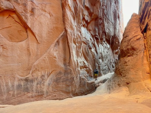 A person walks through a narrow canyon with towering red rock walls and a sandy path.