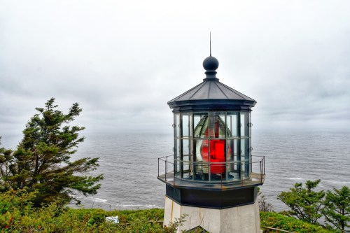 A lighthouse with a red light stands on a cliff overlooking a cloudy ocean. Green trees frame the scene.