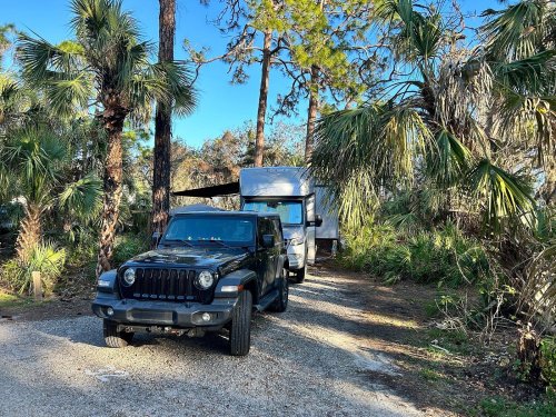 A black Jeep parked on a gravel road surrounded by palm trees and greenery, with a camper trailer behind it.