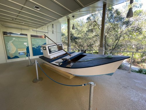 A small boat displayed indoors, surrounded by informational panels and greenery in the background.