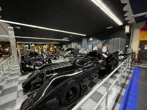 A display of sleek black Batmobiles in a themed exhibit, featuring city skyline murals in the background.