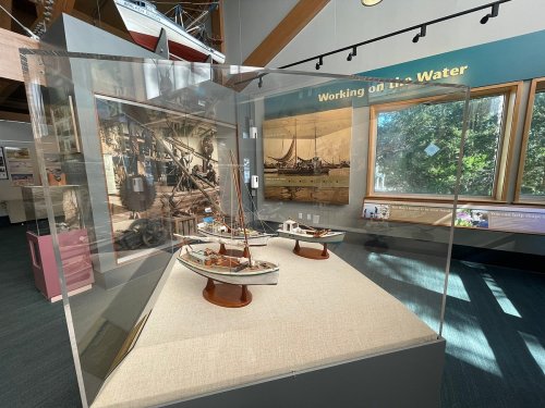 Display of model boats in a glass case, with a backdrop of maritime-themed artwork in a museum setting.