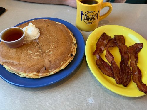 A large pancake with butter and syrup, served with crispy bacon, alongside a cup of coffee in a diner mug.