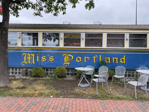 A diner named "Miss Portland" with a blue and yellow exterior, featuring outdoor seating and a cloudy sky.