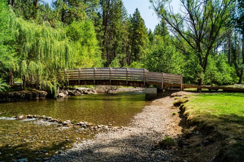 A wooden bridge spans a calm river, surrounded by lush green trees and a rocky shoreline.