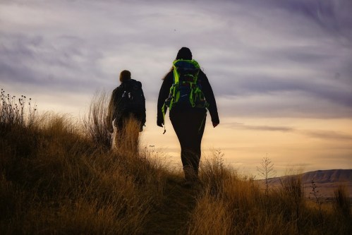 Two hikers with backpacks walking along a grassy trail under a cloudy sky at sunset.