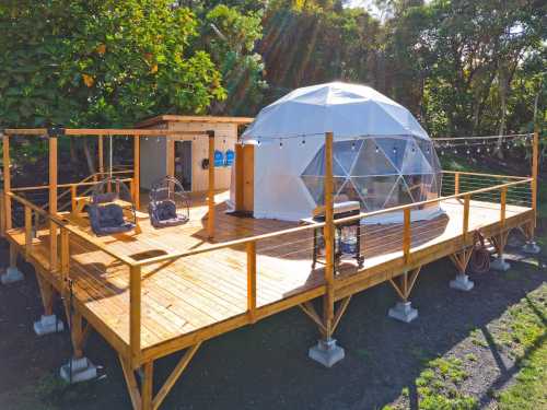 A modern geodesic dome on a wooden deck, surrounded by greenery, with outdoor seating and string lights.