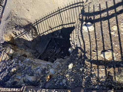 A deep hole in the ground surrounded by rocky terrain, with a shadow of a fence cast over it.