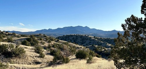 A scenic view of rolling hills and mountains under a clear blue sky, with sparse vegetation in the foreground.