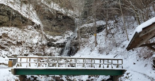 A snowy landscape featuring a wooden bridge in front of a waterfall, surrounded by rocky cliffs and bare trees.