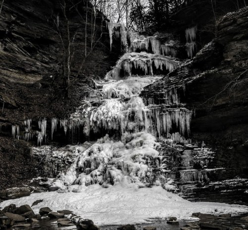 A frozen waterfall cascading down rocky cliffs, surrounded by bare trees and icy formations.